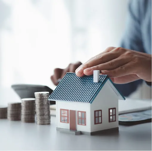 Hands placing a protective gesture over a model house with stacked coins, representing home investment and financial planning